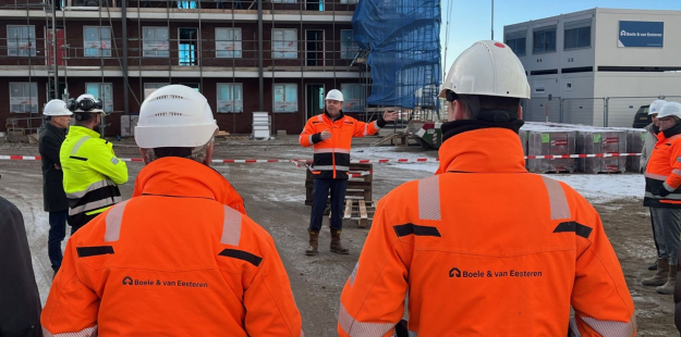 Twee bouwvakkers van achter die luisteren naar hun leidinggevende in de achtergrond 